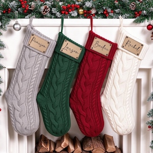 Puede incluir: Cuatro calcetines navideños de punto trenzado en gris claro, verde oscuro, rojo intenso y crema. Cada calcetín tiene una etiqueta rectangular de madera en el puño superior, grabada con los nombres Pearson, Larson, Backer y Rachel en cursiva.