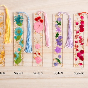 May include: Clear resin bookmarks with dried flowers and gold flakes. Each bookmark has a different flower arrangement and a tassel. Styles include yellow, blue, pink, purple, and red flowers. The bookmarks are rectangular and have the style number at the bottom.