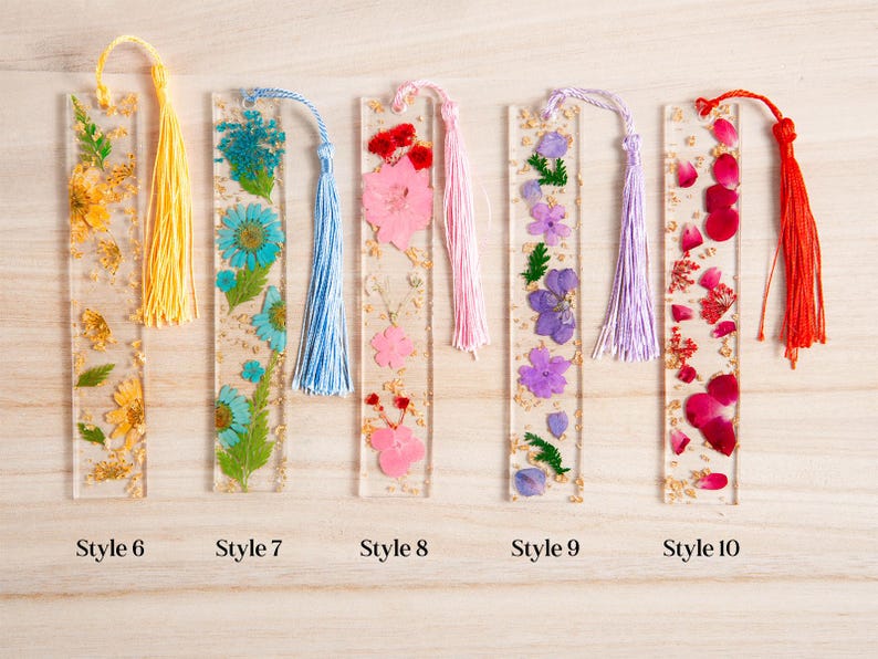 May include: Clear resin bookmarks with dried flowers and gold flakes. Each bookmark has a different flower arrangement and a tassel. Styles include yellow, blue, pink, purple, and red flowers. The bookmarks are rectangular and have the style number at the bottom.