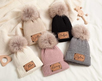 Könnte beinhalten: Sechs Strickmützen in Beige, Schwarz, Weiß, Rosa und Grau, jede mit einem Kunstfell-Bommel. Die Mützen haben ein rechteckiges Lederetikett mit einem Namen. Eine Holzrassel und eine Decke befinden sich im Hintergrund.