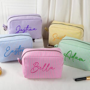 May include: Five personalized cosmetic bags in various colors: purple, yellow, blue, pink, and green. Each bag features a vertical striped pattern and a name embroidered in a matching color. A makeup brush and a small cosmetic container are also visible.