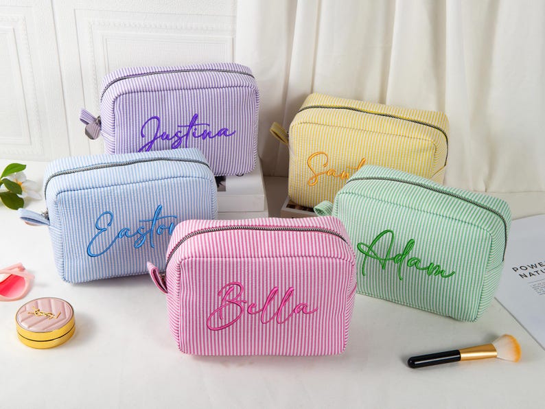 May include: Five personalized cosmetic bags in various colors: purple, yellow, blue, pink, and green. Each bag features a vertical striped pattern and a name embroidered in a matching color. A makeup brush and a small cosmetic container are also visible.