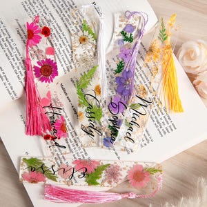May include: Clear resin bookmarks with dried flowers and tassels. Each bookmark features a different flower arrangement and a name in elegant script. The bookmarks are on an open book.