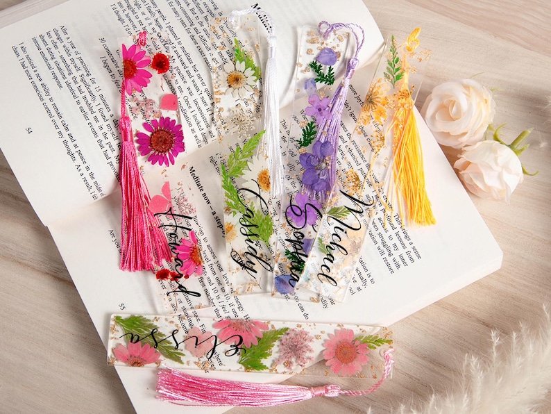 May include: Clear resin bookmarks with dried flowers and tassels. Each bookmark features a different flower arrangement and a name in elegant script. The bookmarks are on an open book.