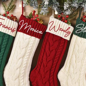 Könnte beinhalten: Vier Weihnachtsstrümpfe aus Zopfstrick in Grün, Creme und Rot, jeder mit einem anderen Namen in Schreibschrift gestickt: Francis, Monica, Wendy und Stanley. Die Strümpfe sind mit Zweigen aus roten Beeren und Grünzeug verziert.
