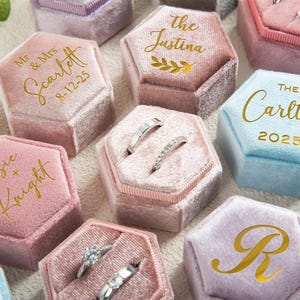 Peut inclure: Plusieurs boîtes à bagues hexagonales en velours de différentes couleurs pastel, notamment rose, bleu et lavande. Certaines boîtes présentent des inscriptions dorées avec des noms et des dates, tandis que d'autres affichent une initiale. Plusieurs boîtes contiennent des bagues en argent.