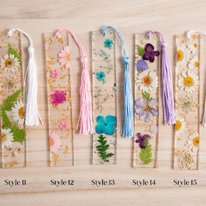 May include: A collection of five clear resin bookmarks, each embedded with dried flowers and gold flakes. Each bookmark has a different color tassel. Styles 11, 12, 13, 14, and 15 are labeled at the bottom.