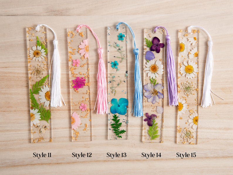 May include: A collection of five clear resin bookmarks, each embedded with dried flowers and gold flakes. Each bookmark has a different color tassel. Styles 11, 12, 13, 14, and 15 are labeled at the bottom.