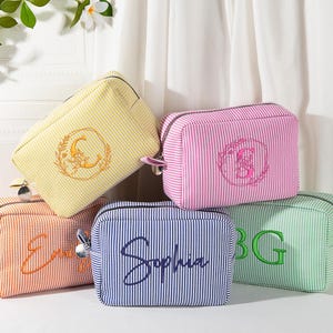 May include: A collection of five striped cosmetic bags in various colors: yellow, pink, orange, blue, and green. Each bag features embroidered details, including initials or a name. The bags have a rectangular shape with a zipper closure, perfect for storing makeup or toiletries.