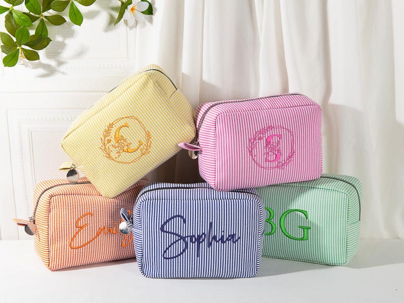 May include: A collection of five striped cosmetic bags in various colors: yellow, pink, orange, blue, and green. Each bag features embroidered details, including initials or a name. The bags have a rectangular shape with a zipper closure, perfect for storing makeup or toiletries.
