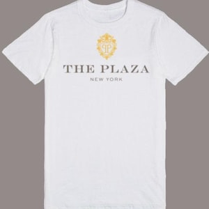 以下が含まれることがあります： 前面に金の紋章と「THE PLAZA NEW YORK」の文字がプリントされた白いTシャツです。