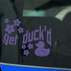 May include: Purple vinyl decal with the phrase "get duck'd", a duck illustration, and floral accents. The decal is designed to be applied to a car window or similar surface.