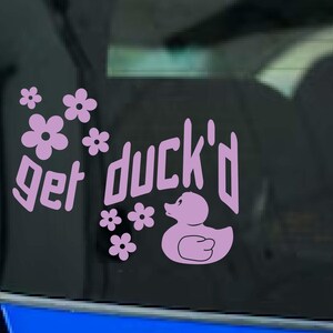 May include: A light purple vinyl decal with the phrase "get duck'd" and a cartoon duck illustration. The decal also features several small flower graphics. The decal is designed to be applied to a car window.