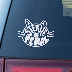 May include: White vinyl decal featuring a cat face design with the words "STERILE & FERAL" in a circular arrangement. The decal is affixed to a dark blue surface, likely a car window.