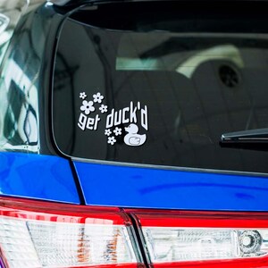 May include: White vinyl car decal with the phrase "get duck'd" and a duck illustration, surrounded by floral accents. The decal is affixed to the rear window of a blue car, near the top.