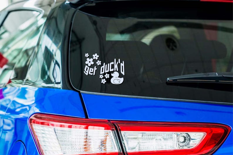 May include: White vinyl car decal with the phrase "get duck'd" and a duck illustration, surrounded by floral accents. The decal is affixed to the rear window of a blue car, near the top.