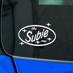 以下が含まれることがあります： 「the Subie crew」の文字がスタイリッシュなフォントで書かれた白い楕円形のステッカー。ステッカーは黒い縁取りと4つのスターバーストのデザインが施されています。ステッカーは車の窓に貼られており、青い車が見えます。