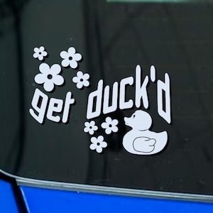 May include: White sticker with the phrase "get duck'd" in a stylized font, accompanied by a cartoon duck and several white flower graphics. The sticker is designed to be applied to a car window or similar surface.