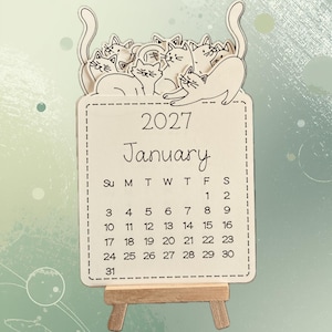 May include: A light-coloured wooden calendar for January 2027, featuring a group of engraved cat illustrations along the top edge. The calendar is displayed on a small wooden easel stand, showing the full month grid with dates from 1 to 31.
