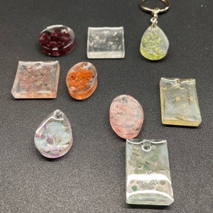 May include: A collection of resin pendants and keychains in various shapes, including round, square, and teardrop. The pendants contain colorful inclusions of red, orange, green, and purple. One keychain features a teardrop pendant with a silver keyring.