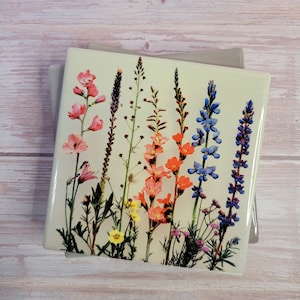 Puede incluir: Un azulejo de cerámica cuadrado con un diseño floral colorido. El azulejo presenta una variedad de flores silvestres en tonos de rosa, naranja, azul y amarillo. Las flores están dispuestas verticalmente sobre un fondo verde claro.