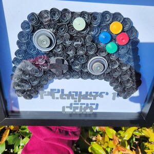 May include: A framed artwork of a black paper-rolled video game controller with colorful buttons. The text "Player 1" is written below the controller.