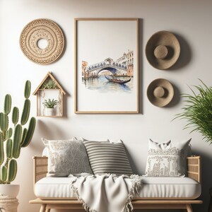 May include: A watercolor painting of a bridge over a canal in Venice, Italy. The painting is framed in a light wood frame and is hanging on a white wall. The wall is decorated with woven wall decor and straw hats.