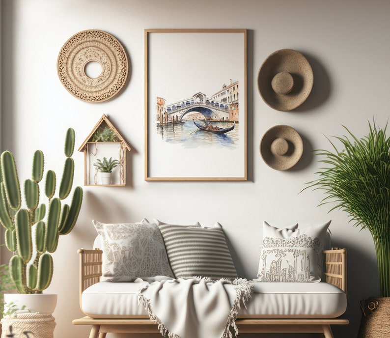 May include: A watercolor painting of a bridge over a canal in Venice, Italy. The painting is framed in a light wood frame and is hanging on a white wall. The wall is decorated with woven wall decor and straw hats.