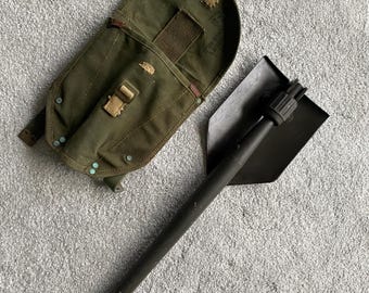 Könnte beinhalten: Ein grüner Canvas-Spatensack mit einer schwarzen Klappschaufel. Die Schaufel hat einen schwarzen Stiel und eine Klinge aus Metall.