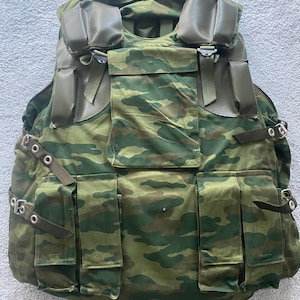 May include: A green and brown camouflage tactical vest with multiple pockets and straps. The vest has a large flap on the front with a zipper and a smaller flap on the back.
