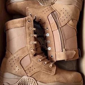 Könnte beinhalten: Ein Paar taktische Stiefel aus beigem Wildleder mit hohem Schaft, seitlichen Reißverschlüssen und Schnürung mit Metallösen. Die Stiefel verfügen über perforierte Seitenteile, eine verstärkte Ferse und eine dicke, profilierte Gummisohle.