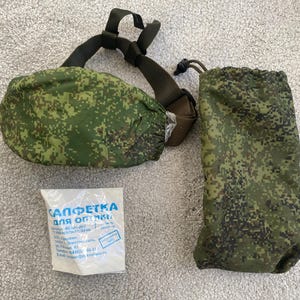 May include: A set of military-style gear in a green and brown pixelated camouflage pattern. The set includes a pouch with a drawstring, a padded case with straps, and a small packet with Cyrillic text.