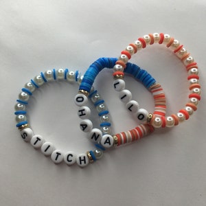 May include: Three beaded bracelets with white beads and colorful bands. The bracelets spell out the names "Stitch", "Lilo", and "Ohana".