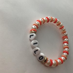 May include: A white beaded bracelet with orange and peach accents. The bracelet spells out the name "LILO" in black letters.