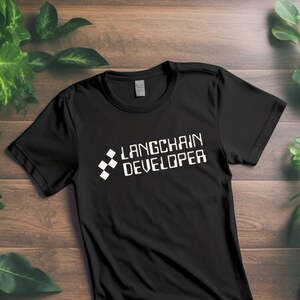 May include: Black t-shirt with the text "Langchain Developer" in white, pixelated font. The shirt has a crew neck and short sleeves. The design includes a geometric pattern on the left side of the text.