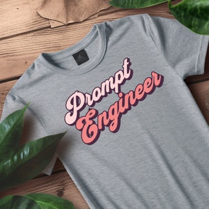 May include: A heather gray crew neck t-shirt with the words "Prompt Engineer" in a retro, layered font. The text is in shades of pink and red, with a dark purple outline. The shirt is displayed on a wooden surface with green leaves.