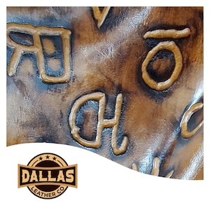 May include: Close-up of a brown leather surface with embossed lettering. The letters are in a lighter brown color, creating a raised effect. The Dallas Leather Co. logo is at the bottom, featuring a shield design with stars and text.