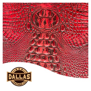 Op de afbeelding: Rode alligatorshuid met een zwart en rood patroon. De huid is getextureerd en heeft een schubbige uitstraling. Het logo van Dallas Leather Co. staat rechtsonder.