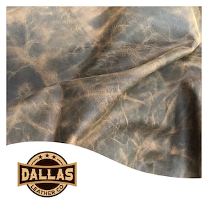May include: Close-up of distressed leather with a blue-gray hue and brown undertones. The leather has a textured appearance with visible grain and folds. The Dallas Leather Co. logo is in the lower left corner.