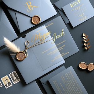 Könnte beinhalten: Ein Set aus staubblauen Hochzeitspapeterie-Artikeln, bestehend aus einem Umschlag mit goldenem Wachssiegel, einer RSVP-Karte und einer Einladung mit goldenem Schriftzug Megan und Jack, verziert mit Kordel und einer weißen Feder sowie Briefmarken.