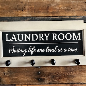 May include: A white and black wooden sign with the text "Laundry Room" and "Sorting life one load at a time." The sign has four black metal hooks on the bottom.