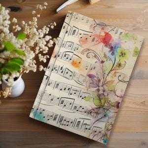 May include: A vintage-style notebook with a watercolour floral design on a background of sheet music. The flowers are in shades of pink, blue, and yellow, and the music notes are black. The notebook has a cream-coloured cover.