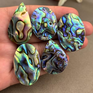 May include: Five iridescent abalone shell cabochons in different shapes. The cabochons show a vibrant mix of blues, greens, and golds, with unique patterns. The shapes include teardrop and oval forms, ideal for jewellery making or crafts.