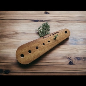 May include: A wooden kitchen tool with five holes and the text "Wood and Splinter" engraved on it. The tool is on a wooden surface with green herbs.