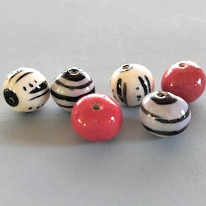 May include: A collection of ceramic beads in various colors and patterns. The beads include white with black stripes and abstract designs, and solid red beads. Each bead has a hole for stringing.