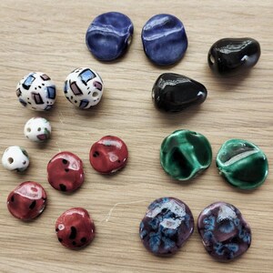 May include: A selection of ceramic beads in diverse shapes and hues. The beads feature shades of blue, black, red, green, and white, with patterned and solid finishes. Some are spherical, while others have irregular forms.