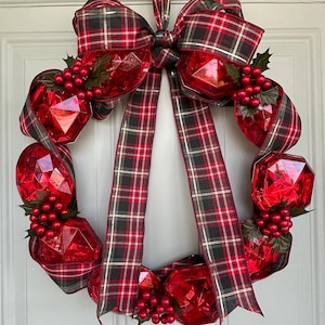 Puede incluir: Una corona navideña festiva con grandes adornos rojos facetados y bayas rojas. La corona está adornada con una cinta a cuadros roja, negra y dorada. Una pieza decorativa para la temporada navideña.