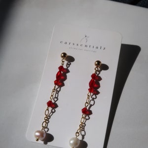 May include: A pair of gold earrings with red crystal beads and a white pearl at the end of each earring. The earrings are on a white card with the text "earsssentialz whimsical earrings".