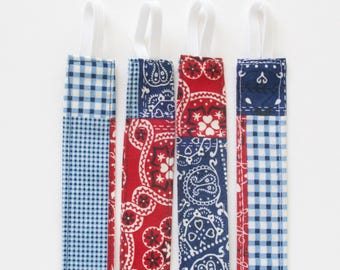 Puede incluir: Cuatro marcadores de tela con lazos elásticos blancos. Los marcadores presentan un diseño de patchwork con cuadros azules, paisley rojo y patrones paisley azules. Los marcadores están dispuestos verticalmente sobre un fondo blanco.