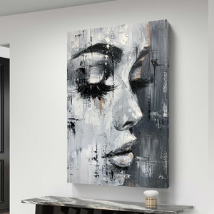 May include: Abstract oil painting of a woman's face in profile, in grey, black, and white with gold accents. The artwork has thick brushstrokes and a textured look. The painting is displayed above a modern black table.